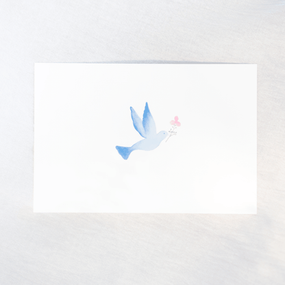 A white rectangular card featuring a delicate watercolor illustration of a blue bird holding a small pink flower, photographed on a light neutral background.