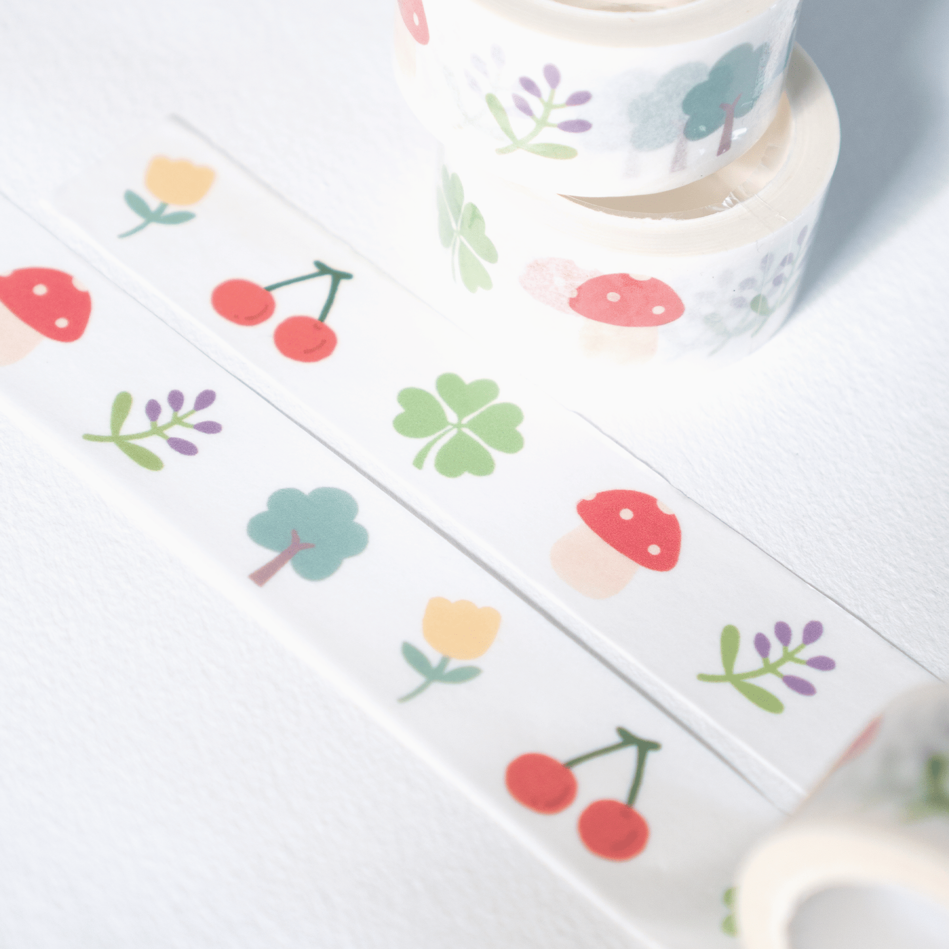 White washi tape with a colorful spring and summer pattern, including cherries, flowers, and mushrooms, shown in rolls and a flat strip on a white background.